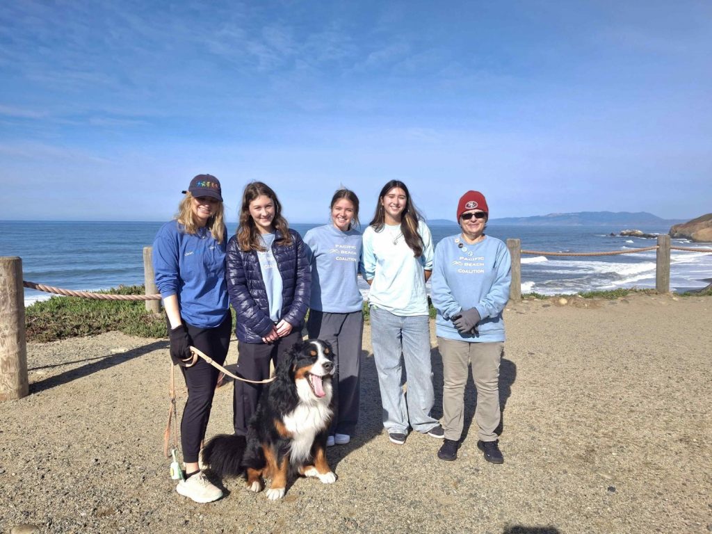 Four Pacific Beach Coalition volunteers, a dog and a site captain pose with the ocean in the background