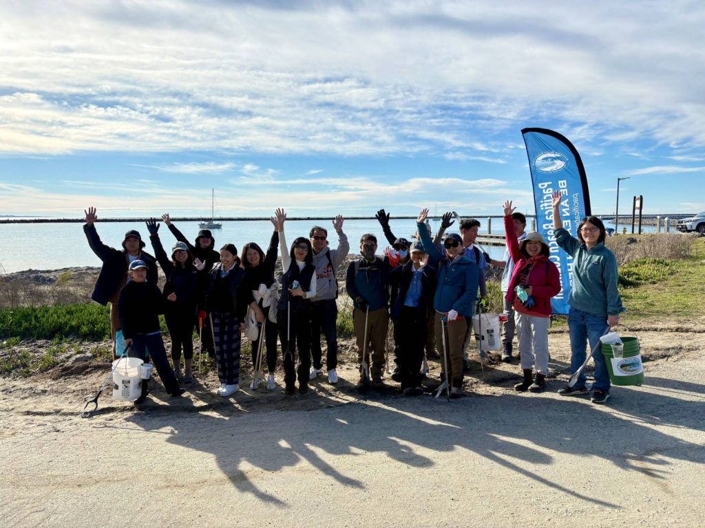 A group of volunteers raising their hands stand with trash pickers and buckets in front of a harbor