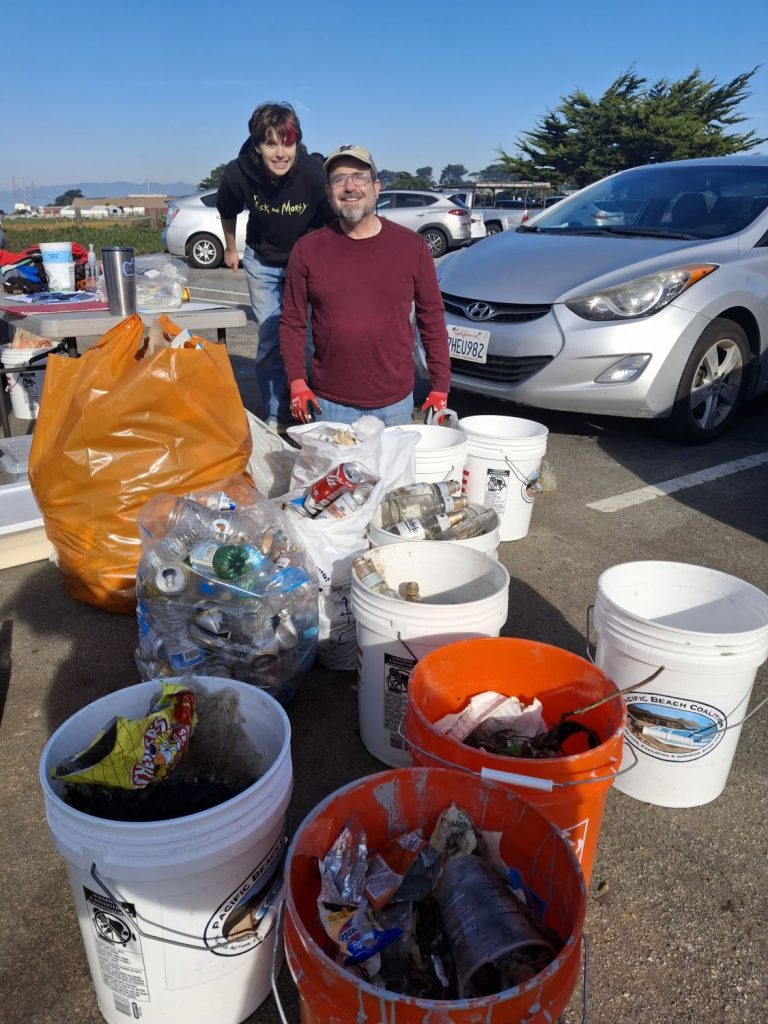 Two volunteers pose with buckets filled with recyclables and trash picked up during a beach cleanup