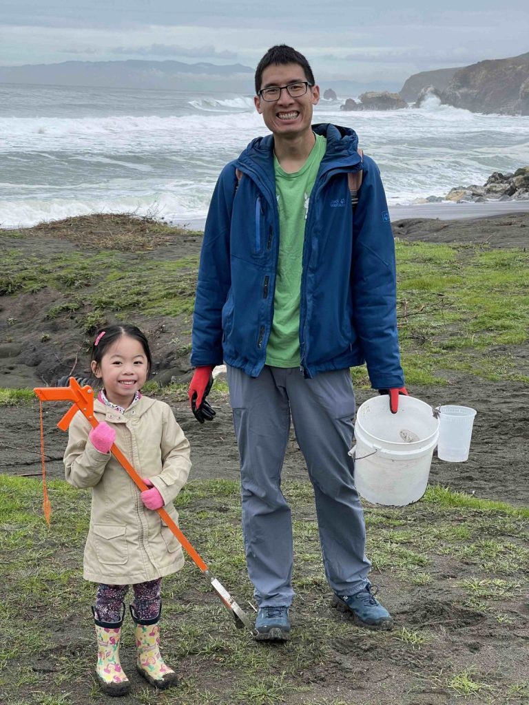 A child and adult pose with their trash-collecting tools at a beach cleanup