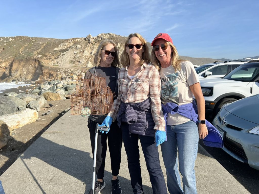 Three women standing near the beach; one is holding a birdcage found during a beach cleanup