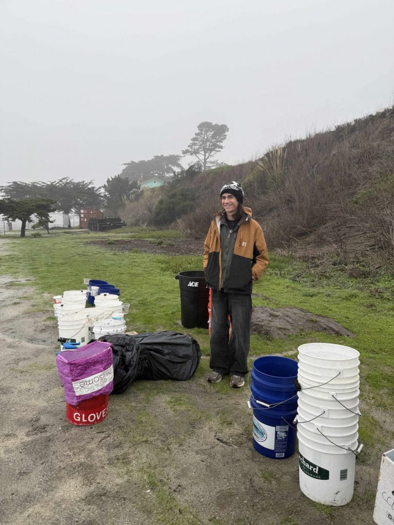 A young adult stands near buckets that will be used for a beach cleanup