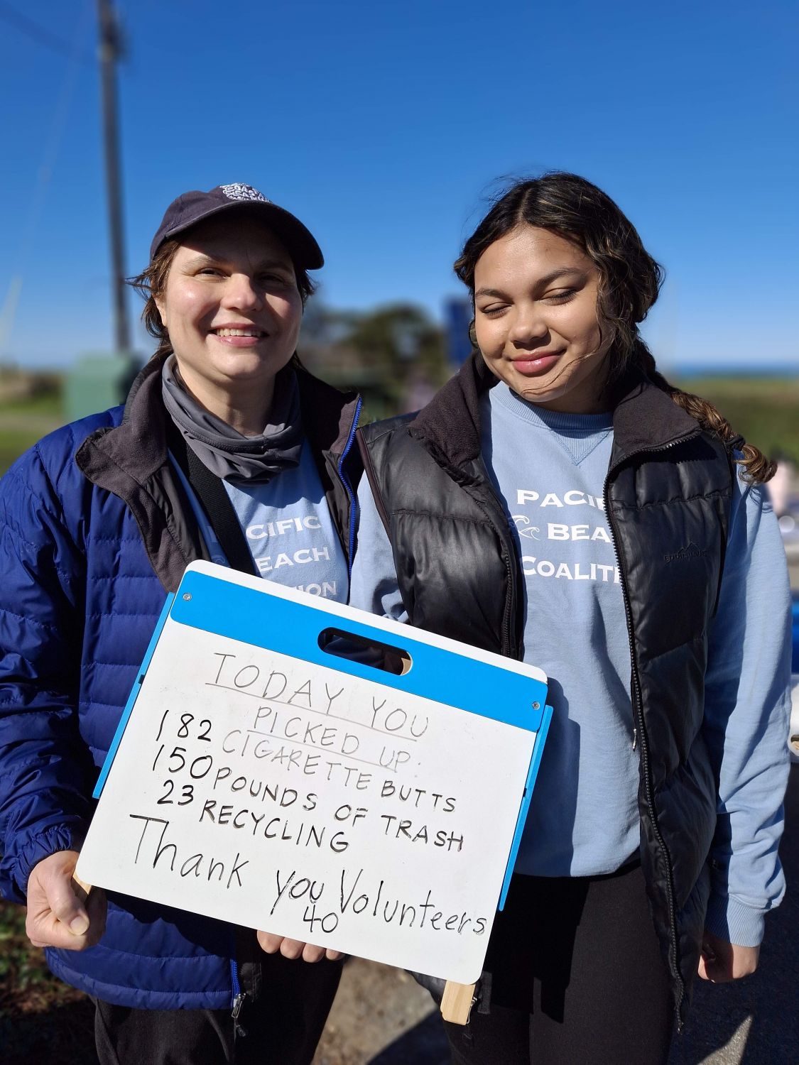 Two female volunteers pose with a poster that displays results from a February 2025 PBC cleanup